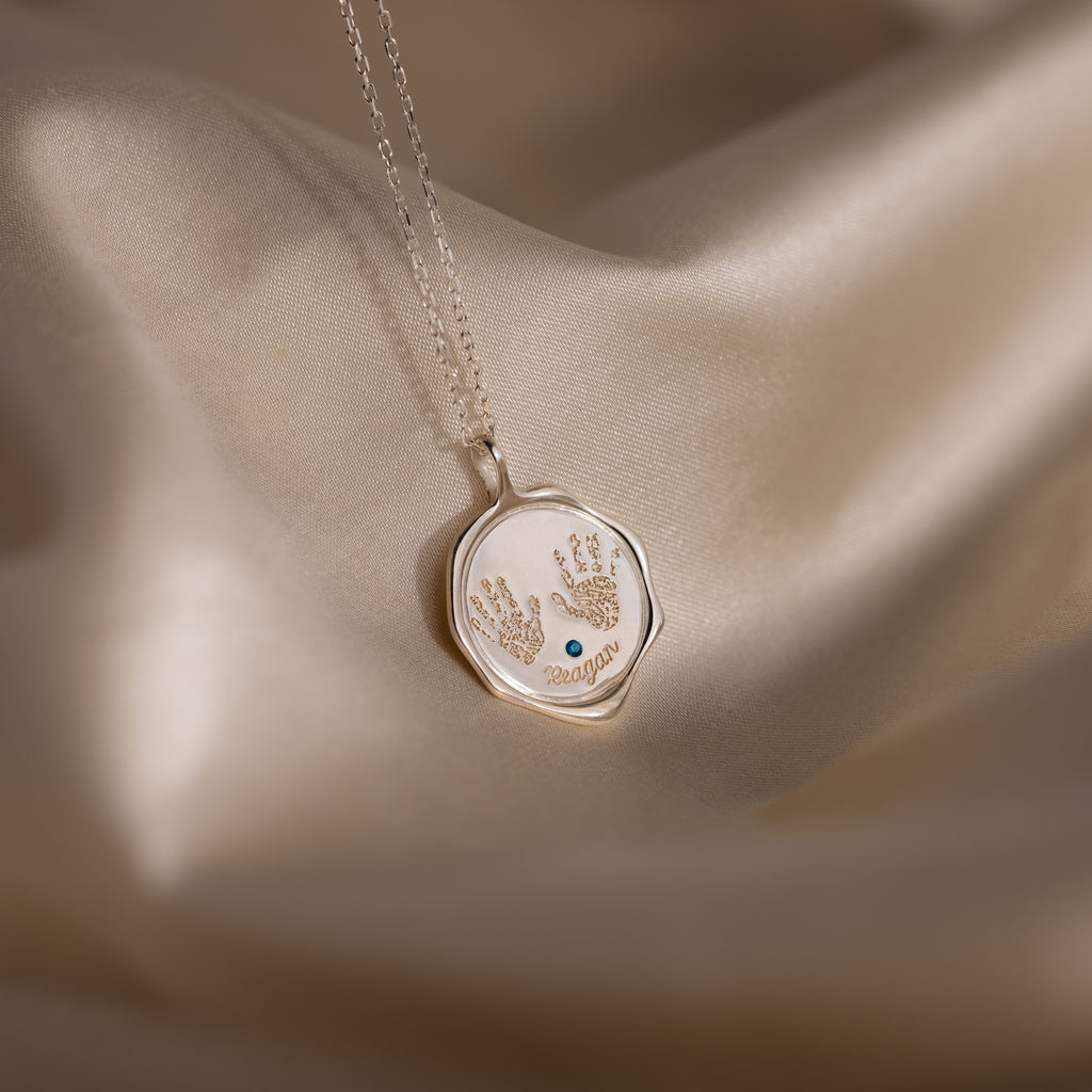 The Baby Hand & Footprint Necklace in Sterling Silver features a pendant with two tiny handprints and the name Reagan, beautifully displayed on silky beige fabric—an ideal piece of personalized baby jewelry.
