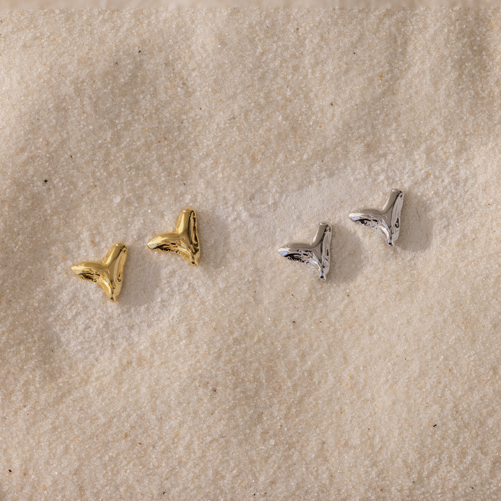 Two pairs of small, metallic Shark Tooth Flatback Studs—one gold, one silver—resting on a sandy surface make the perfect beach accessory.