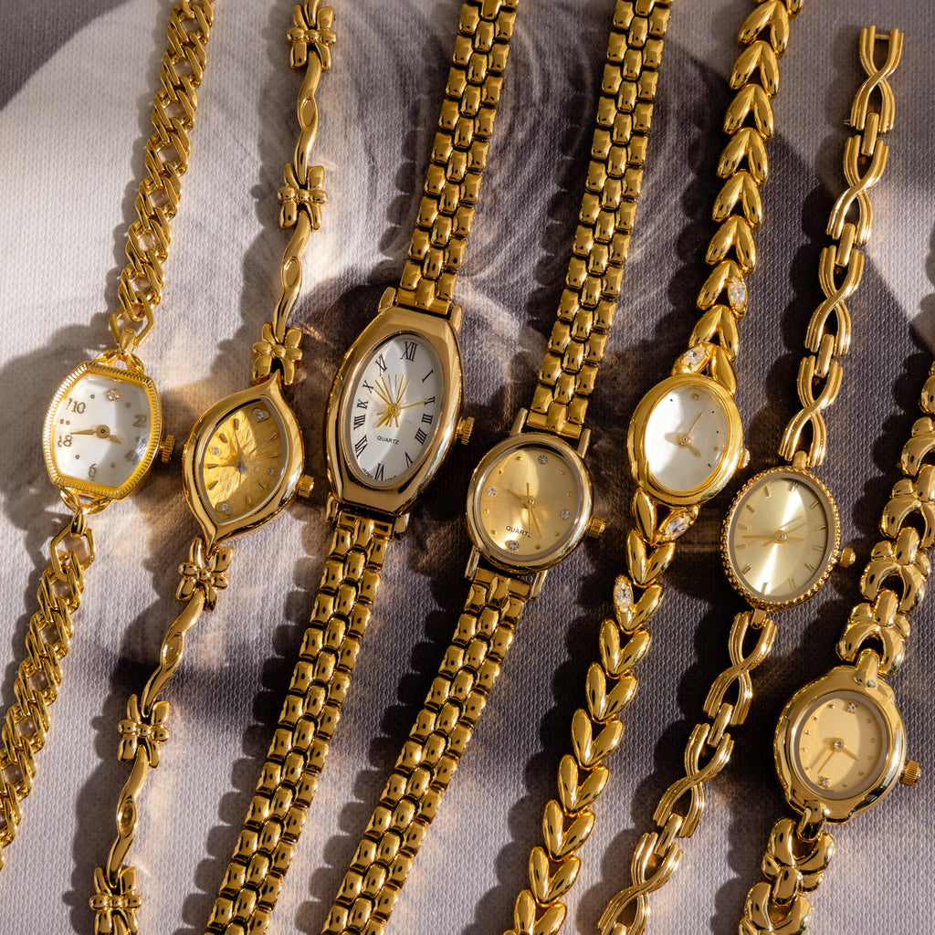 Seven Heirloom Chain Link Timepieces, featuring various designs like diamond accents and mother of pearl dials, are arranged side by side on a patterned background.