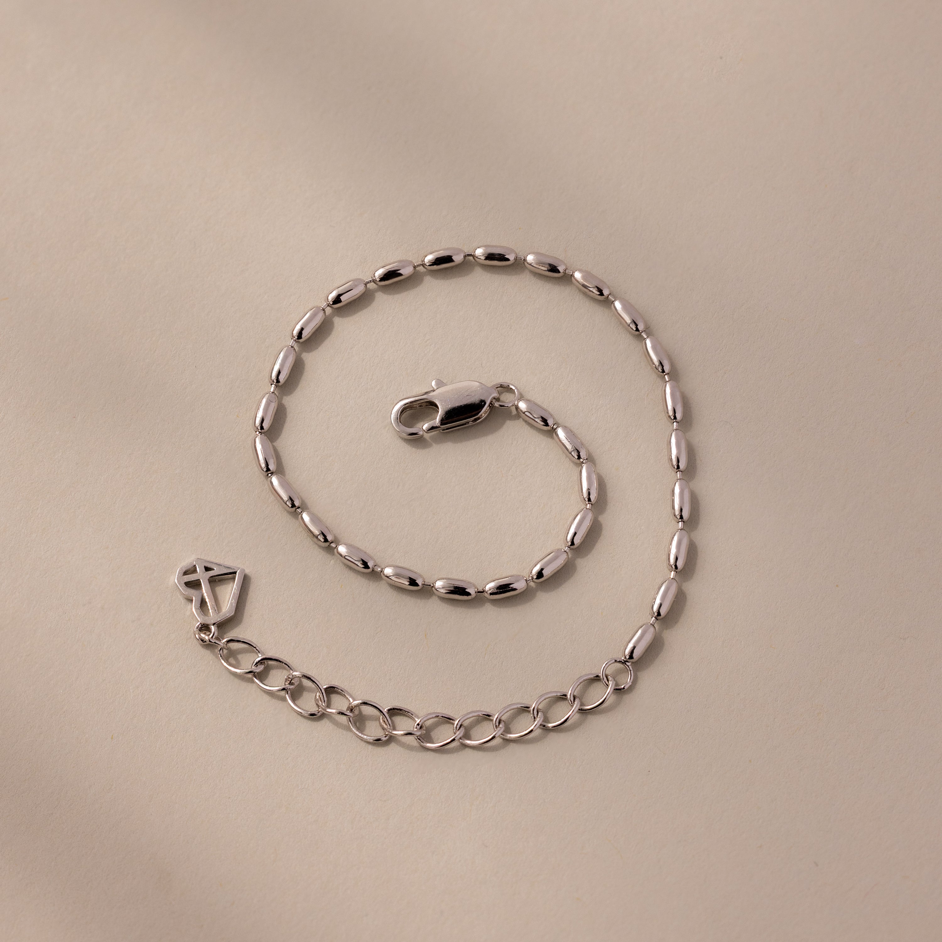 The Oval Beaded Bracelet features silver oval links and polished beads with a lobster clasp, displayed on a beige surface—ideal for fans of modern classic jewelry.