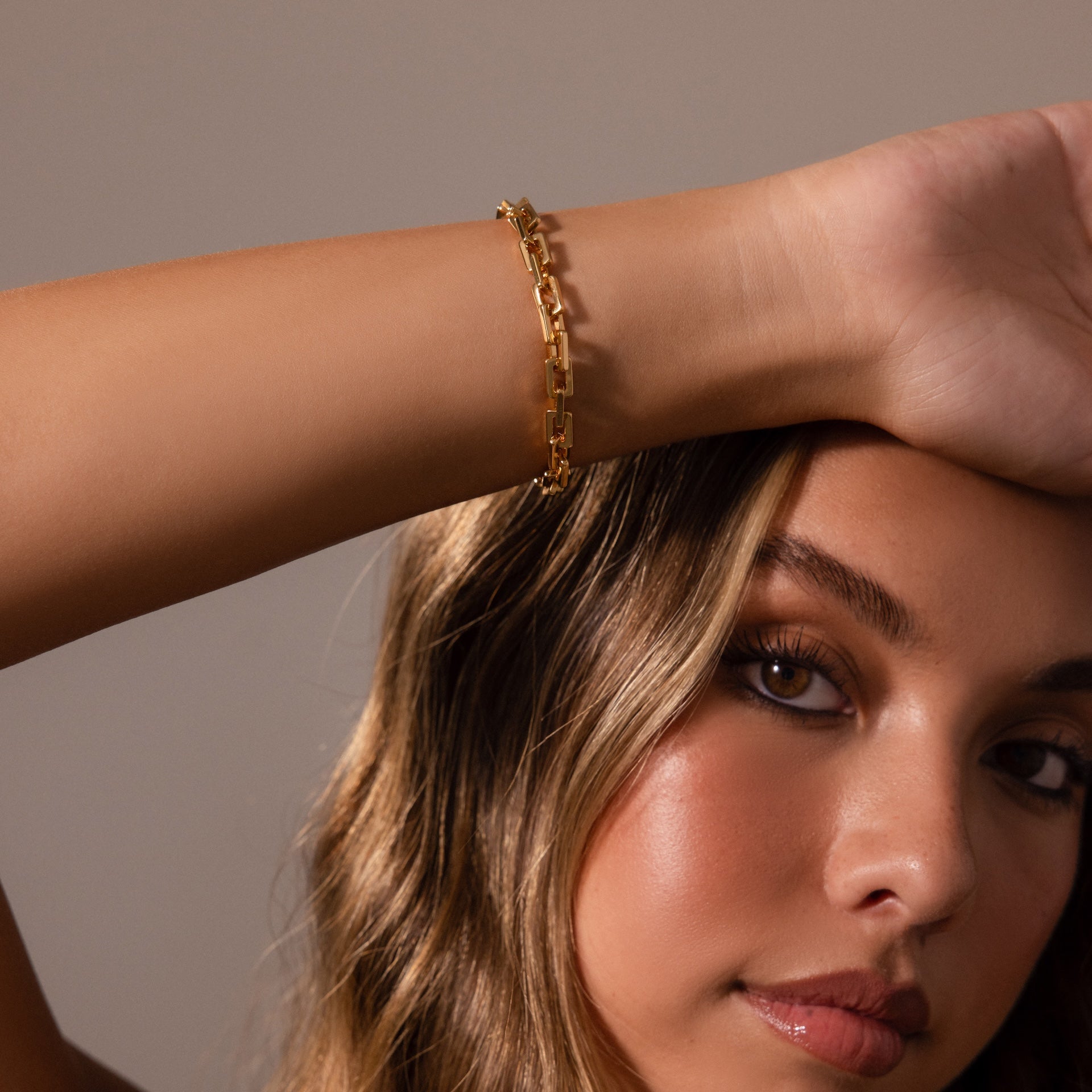 A woman with long brown hair wears the Eternal Link Bracelet in 18K Gold, featuring a subtle geometric design, as she rests her arm on her forehead and looks at the camera.