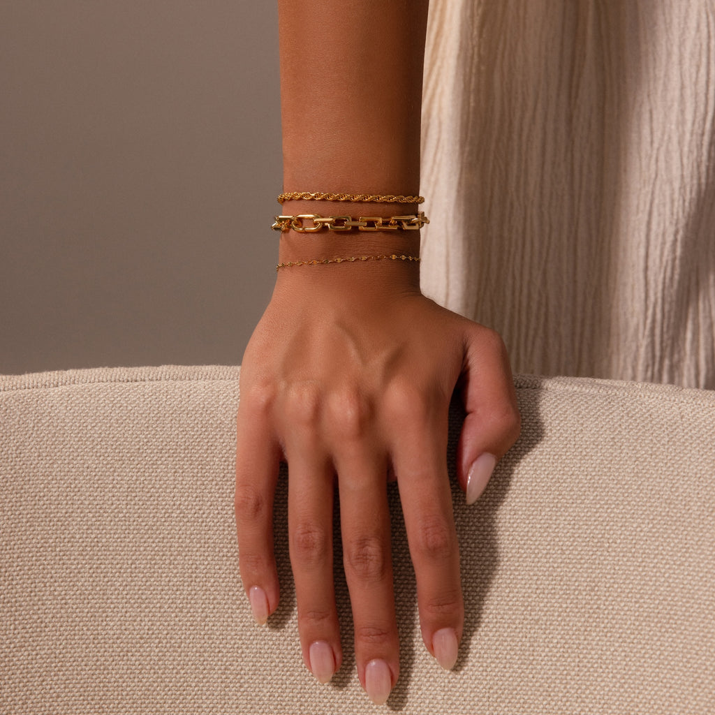 A hand with nude nails wears the Eternal Link Bracelet and two other gold bracelets on a beige textured surface—a modern essentials showcase.