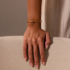A hand with nude nails wears the Eternal Link Bracelet and two other gold bracelets on a beige textured surface—a modern essentials showcase.