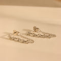 The Diamond Chain Drop Earrings in Silver, featuring five clear gemstones each, are elegantly displayed on a light beige surface.