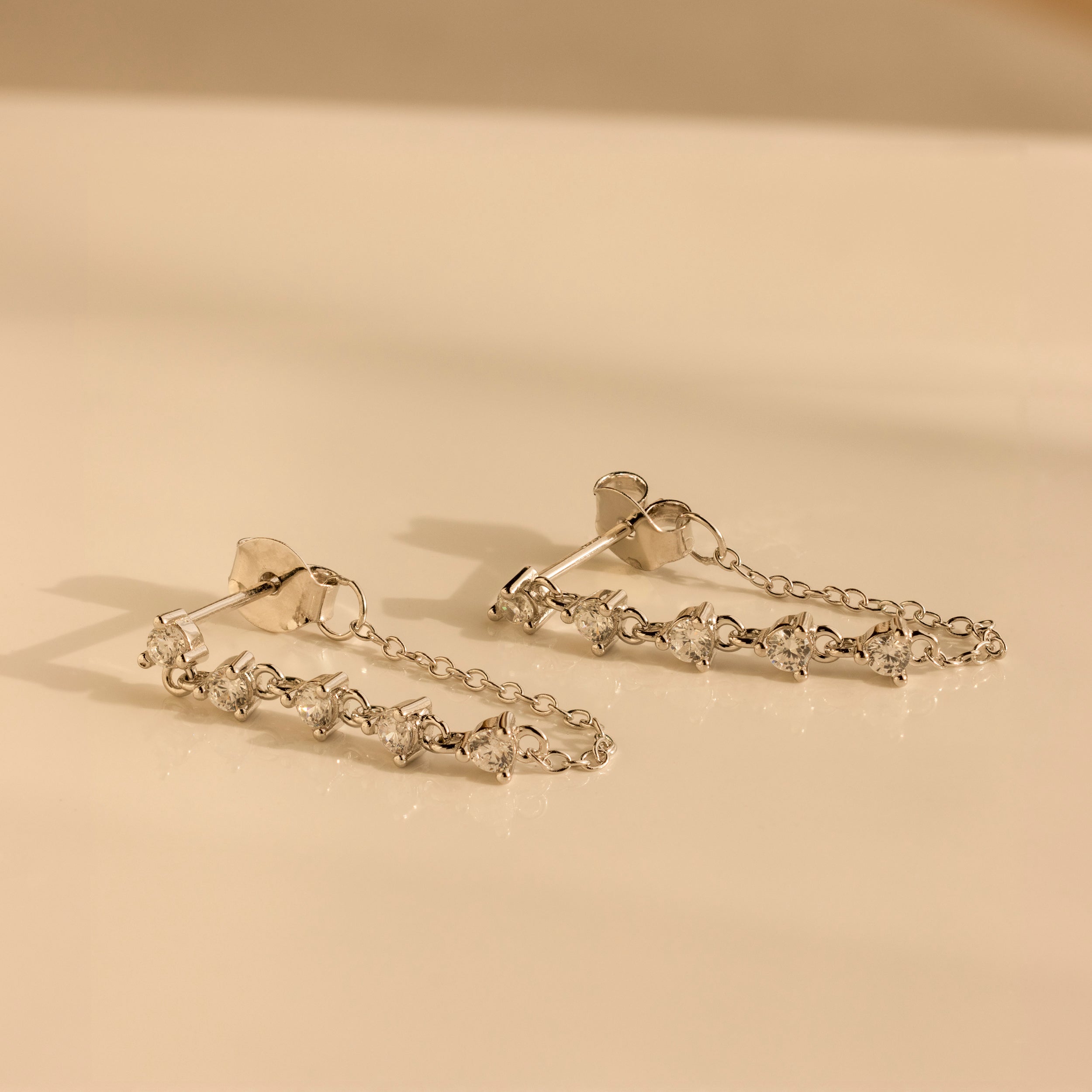 The Diamond Chain Drop Earrings in Silver, featuring five clear gemstones each, are elegantly displayed on a light beige surface.