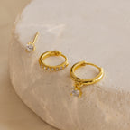 The Classic Diamond Earrings Set features three gold earrings with clear stones, including a classic stud design, displayed on a round beige textured surface.