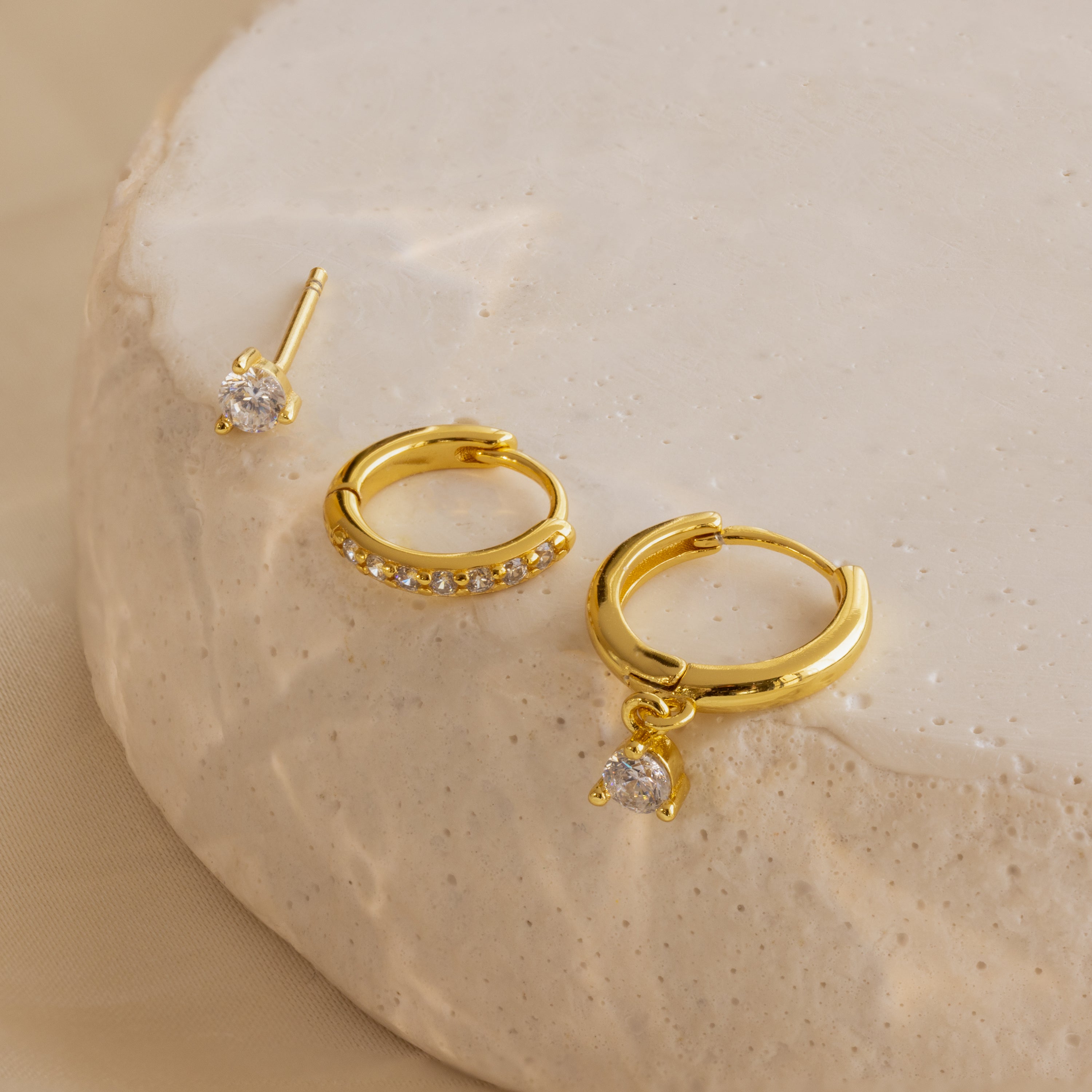 The Classic Diamond Earrings Set features three gold earrings with clear stones, including a classic stud design, displayed on a round beige textured surface.