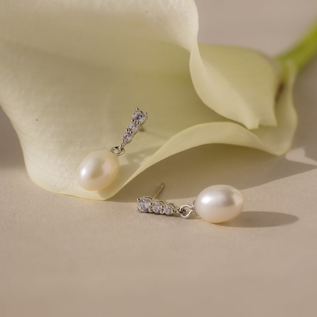 The Diamond Pearl Droplet Earrings in Sterling Silver are displayed beside a white calla lily on a beige surface.