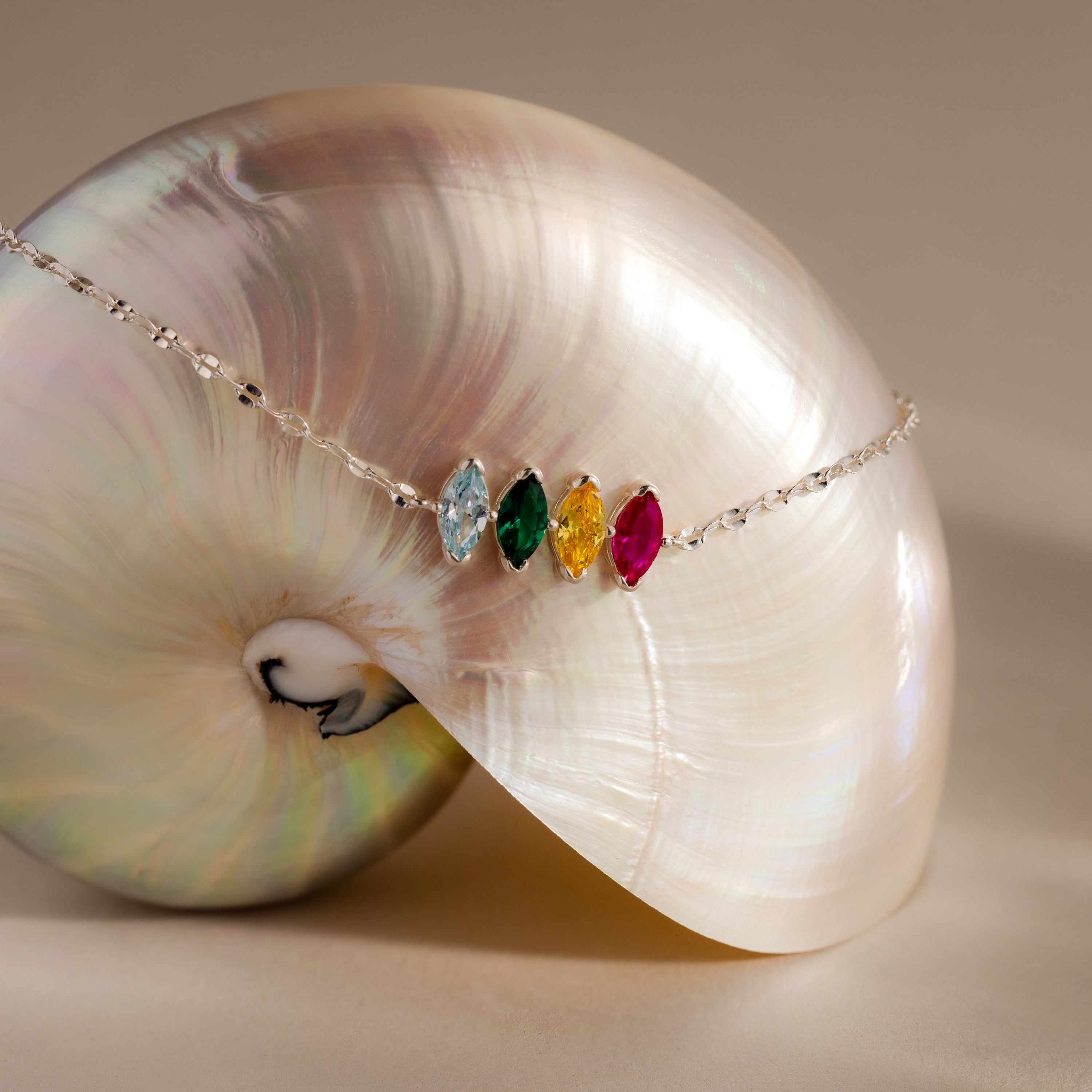 The Multiple Marquise Birthstone Bracelet is a customizable silver piece featuring four vibrant gemstones atop a shiny iridescent seashell—a striking addition to birthstone jewelry.