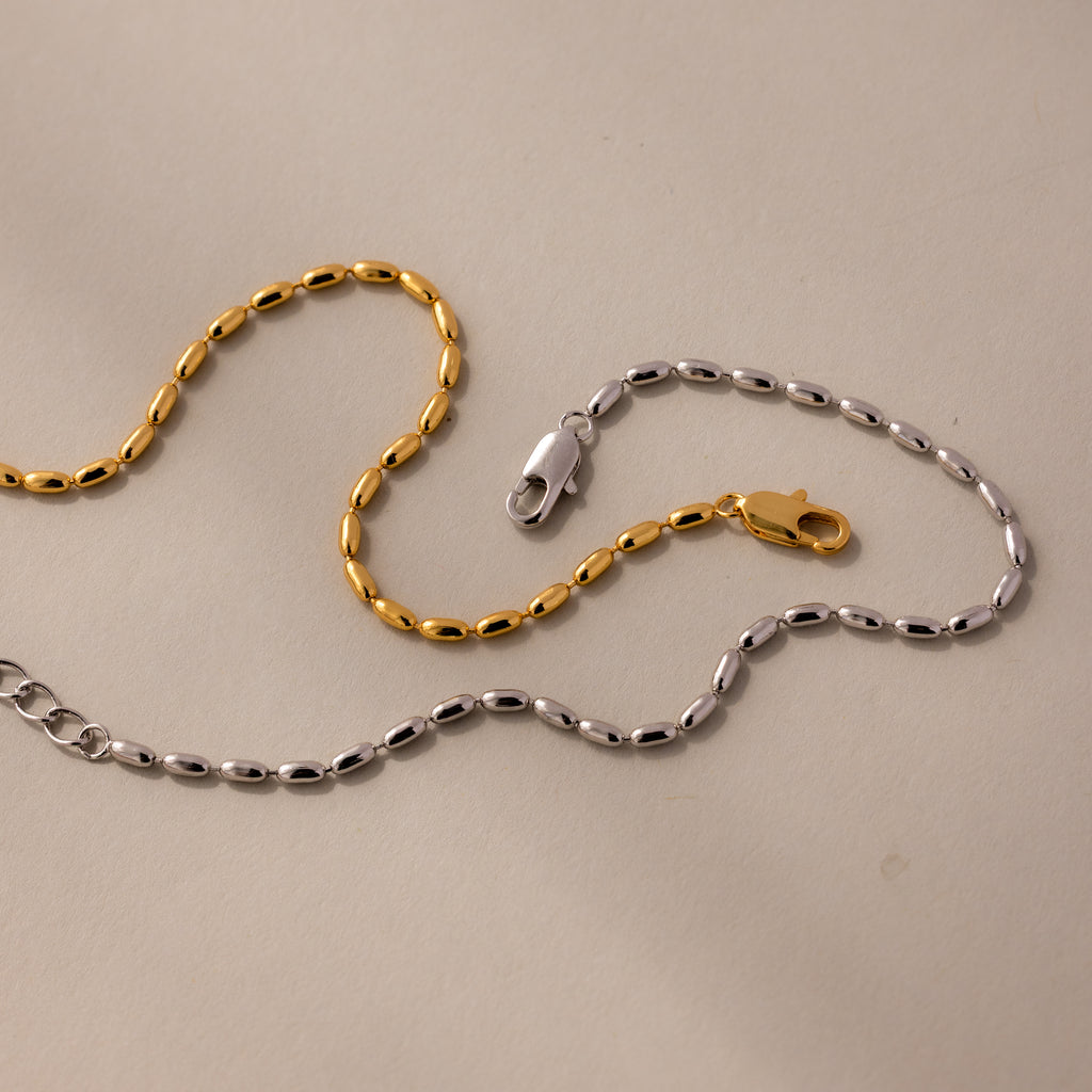 The Oval Beaded Bracelet features polished gold and silver beads with clasps, displayed on a light beige surface for a modern classic jewelry style.
