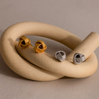 Rope Knot Studs in gold and silver, textured and stylish, are elegantly showcased on a smooth beige ceramic loop—blending timeless design with modern flair.