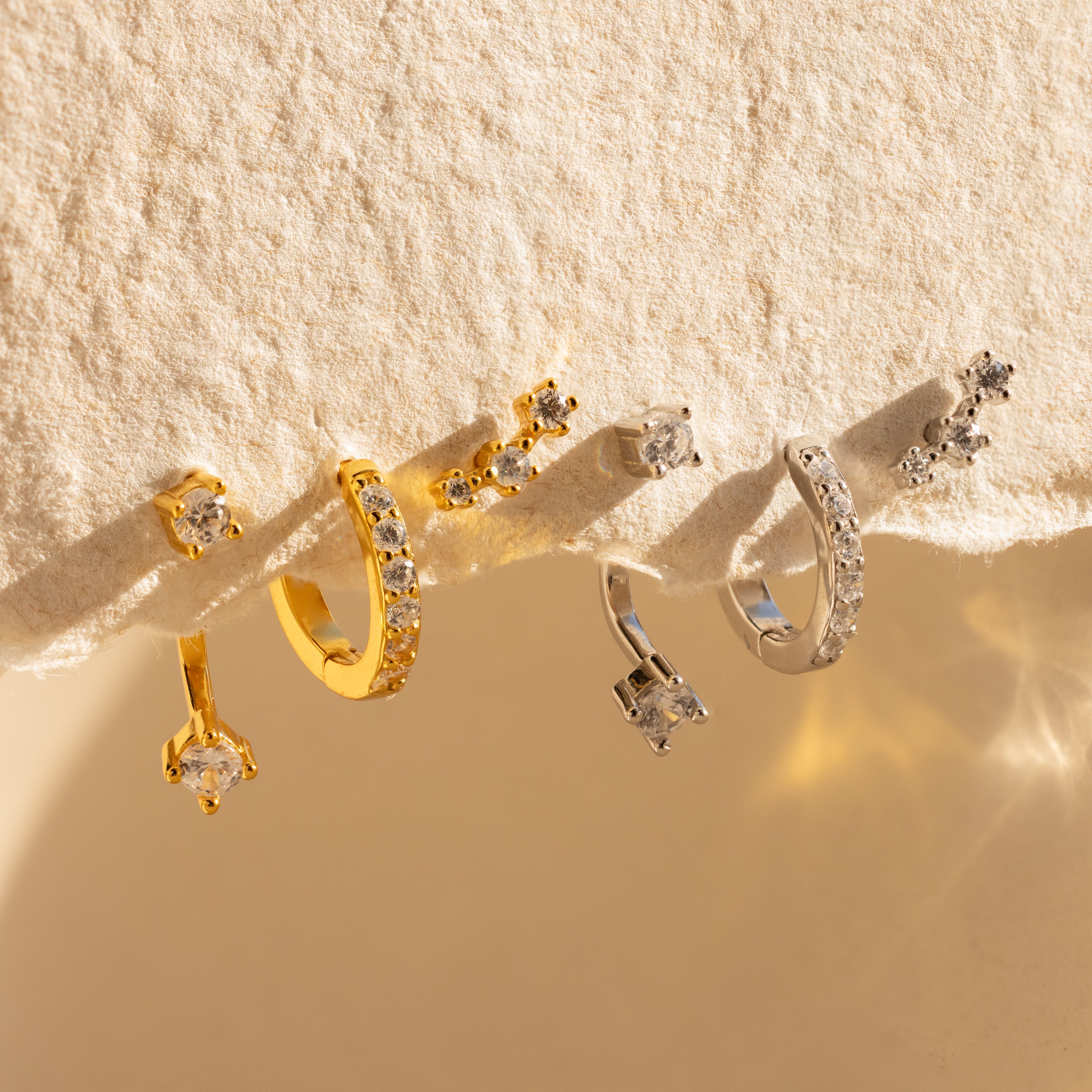 Diamond Earrings Set displayed on a textured beige surface in sunlight, featuring gold and silver Pave Huggie Hoops adorned with small gemstones.