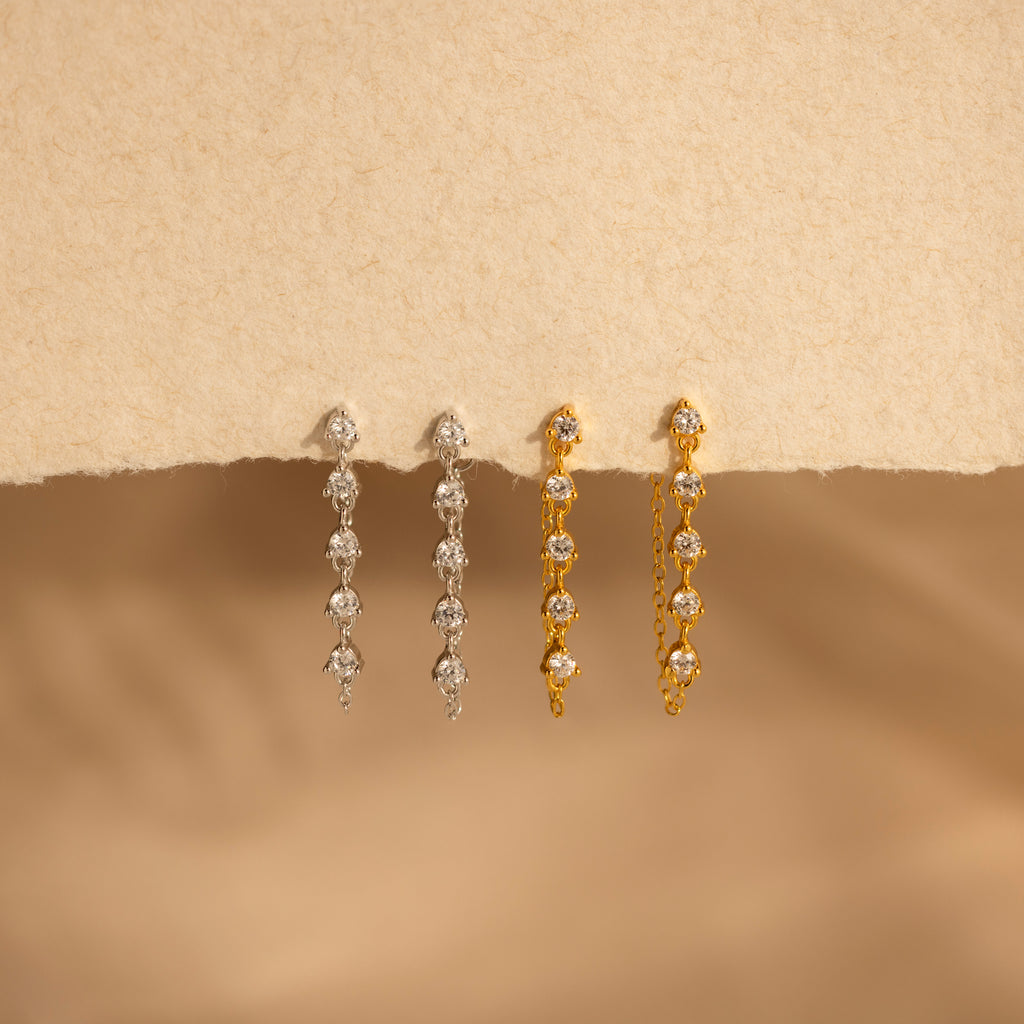 The Diamond Chain Drop Earrings in 18K Gold are displayed on a textured beige surface, highlighting their elegant design.