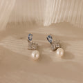 The Aquamarine Pearl Drop Earrings in Silver, featuring blue gemstones and silver accents, exude vintage glamour as they rest elegantly on a soft beige surface.
