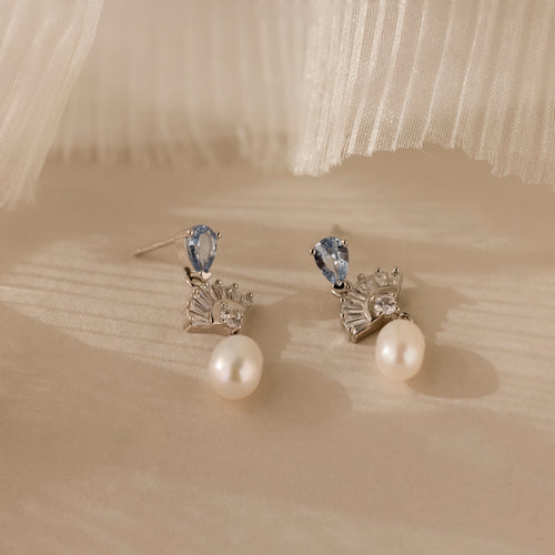 The Aquamarine Pearl Drop Earrings in Silver, featuring blue gemstones and silver accents, exude vintage glamour as they rest elegantly on a soft beige surface.
