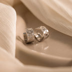 Two Pave Checkered Huggies in Silver with square gem details rest on a soft beige fabric background.