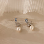 The Aquamarine Pearl Drop Earrings feature blue gemstones, clear crystals, and round white pearls for vintage glamour, beautifully displayed on a soft beige surface.