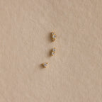 Three Tiny Diamond Flatback Studs Set in 18K Gold are arranged vertically on a textured beige surface.