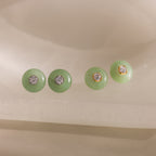 Close-up of two pairs of round green jade stud earrings in gold and silver, each featuring a sparkling CZ diamond center, displayed on a neutral background.