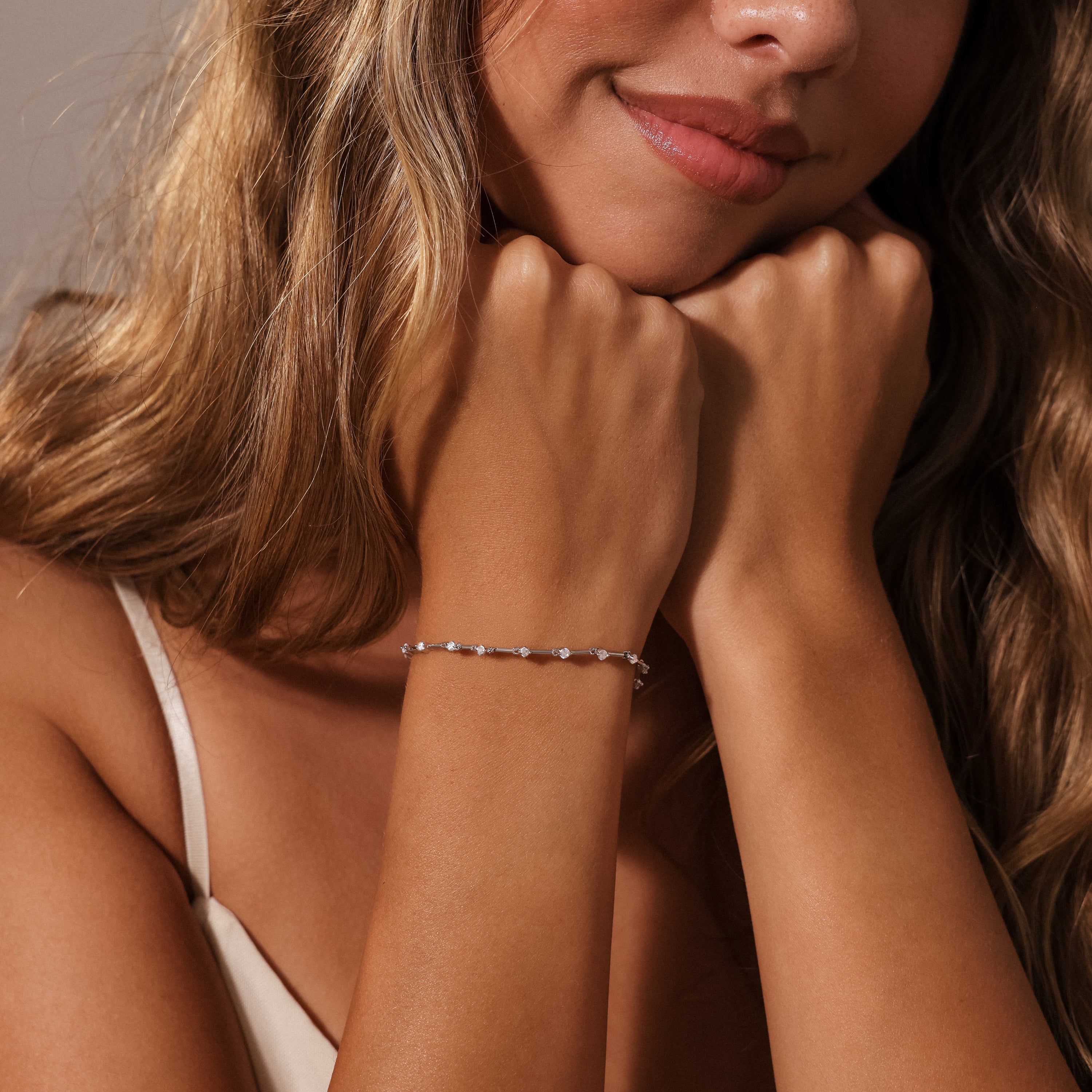 A woman with wavy hair smiles softly, her hands near her chin, wearing the Sprinkle Diamond Tennis Bracelet in Silver.