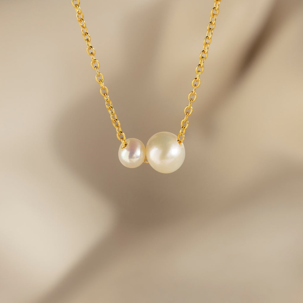 The Dainty Duo Pearl Necklace features two white pearls of varying sizes on a gold chain, perfect as elegant wedding jewelry, displayed against a soft beige blurred background.