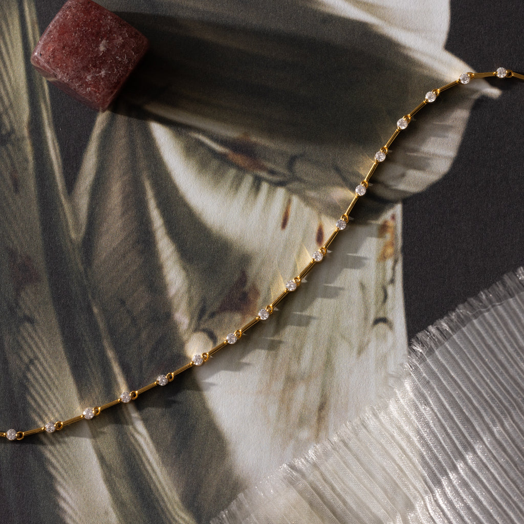 The Sprinkle Diamond Tennis Bracelet—a delicate gold chain with small gems—rests on fabric near a reddish stone and pleated edge, perfect for a dainty look.