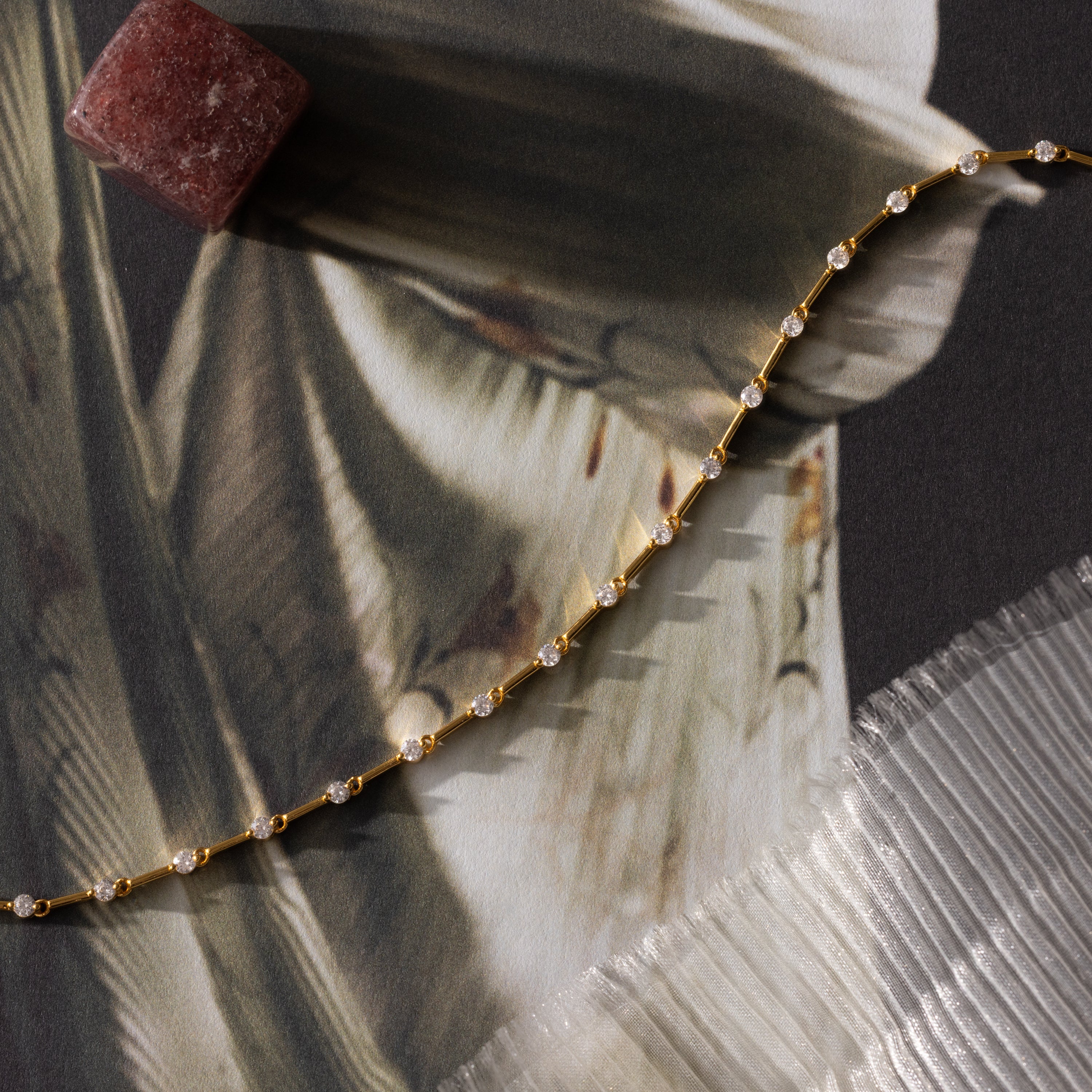 The Sprinkle Diamond Tennis Bracelet—a delicate gold chain with small gems—rests on fabric near a reddish stone and pleated edge, perfect for a dainty look.
