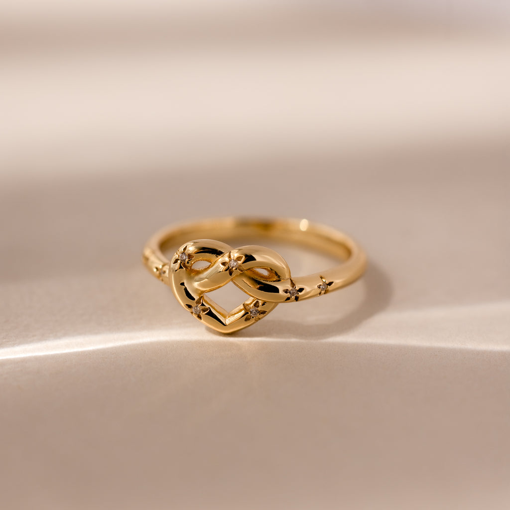 The Starry Love Knot Ring, featuring a delicate gold band with an intricate love knot design, rests on a soft beige surface—symbolizing timeless elegance and heartfelt connection.