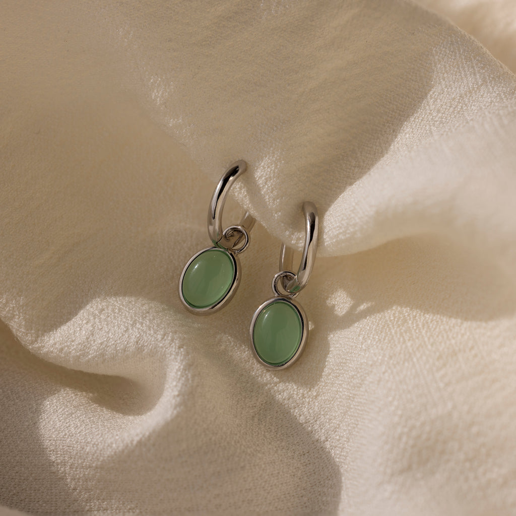Jade Drop Hoops in Silver with oval jade gemstones rest on a soft, cream-colored textured fabric.