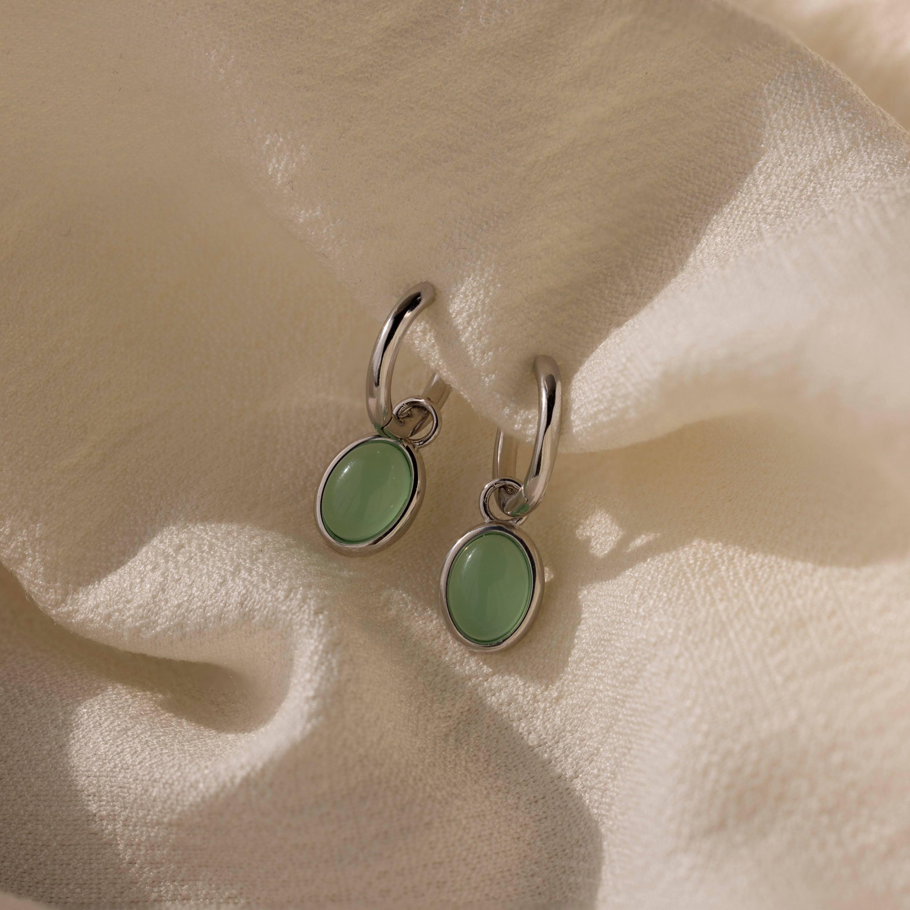 Jade Drop Hoops in Silver with oval jade gemstones rest on a soft, cream-colored textured fabric.