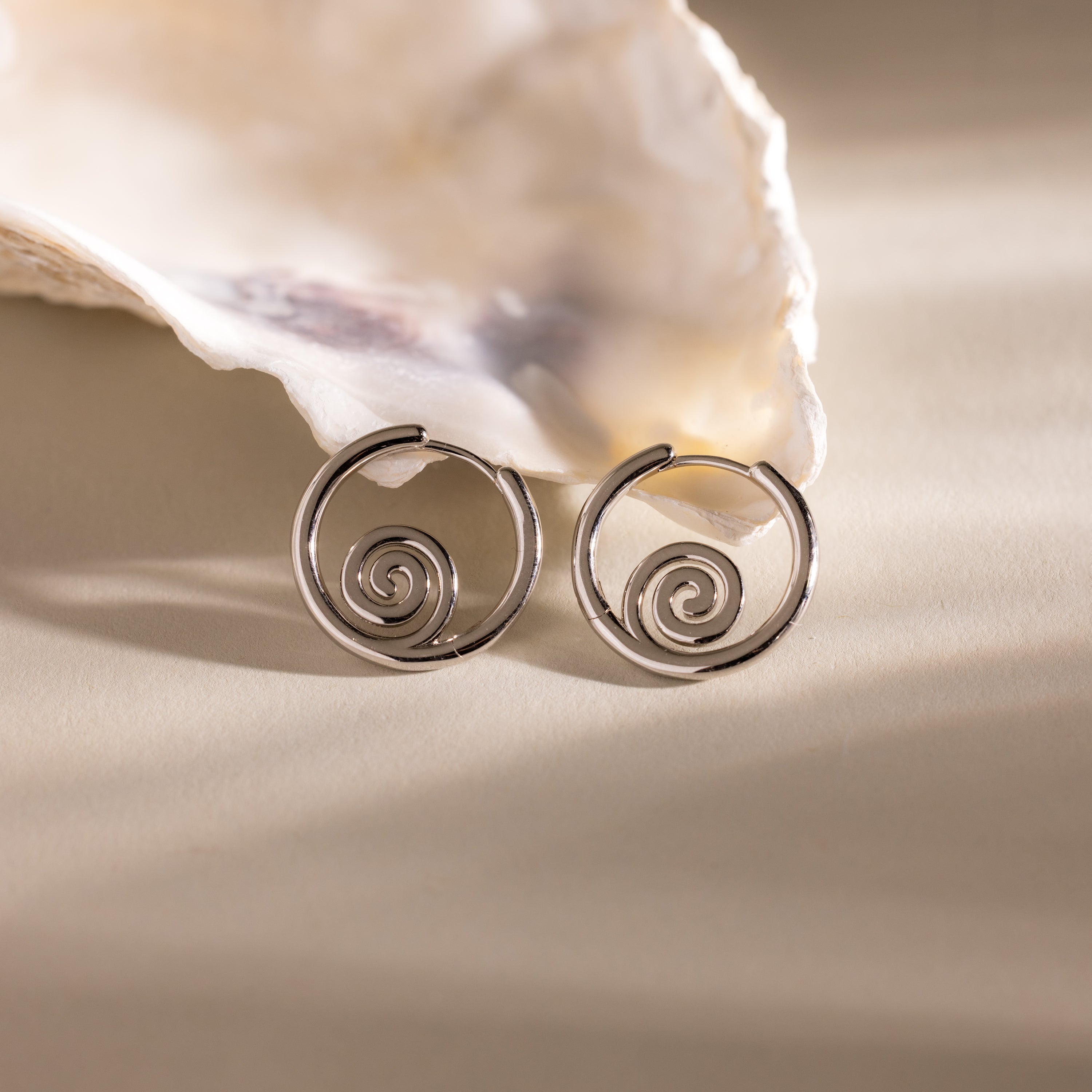 Spiral Swirl Hoops in Silver displayed before a white seashell on a beige background—ideal vacation jewelry or for giving your outfit a boho, beachy vibe.