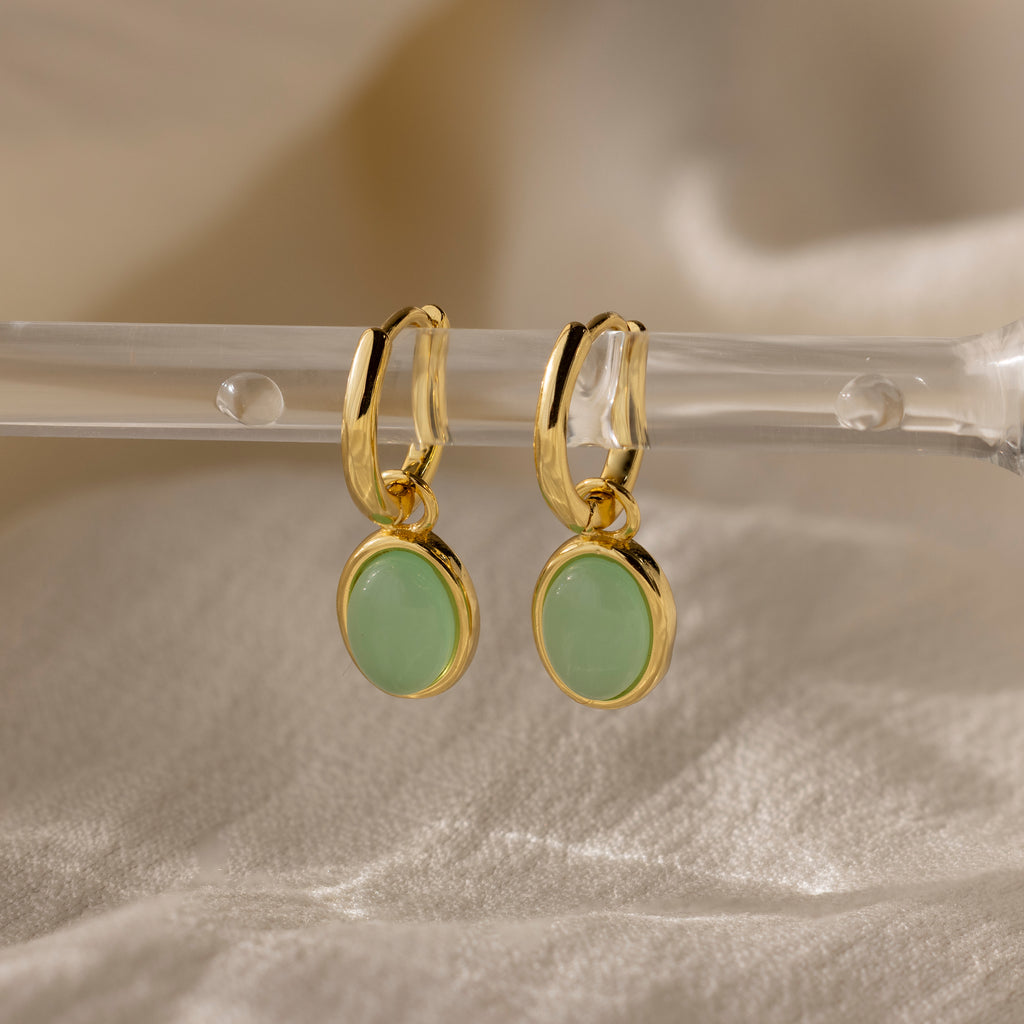 Jade Drop Hoops in 18K Gold with round jade gemstone accents hang from a clear rod on soft beige fabric, combining classic huggie style with vibrant elegance.