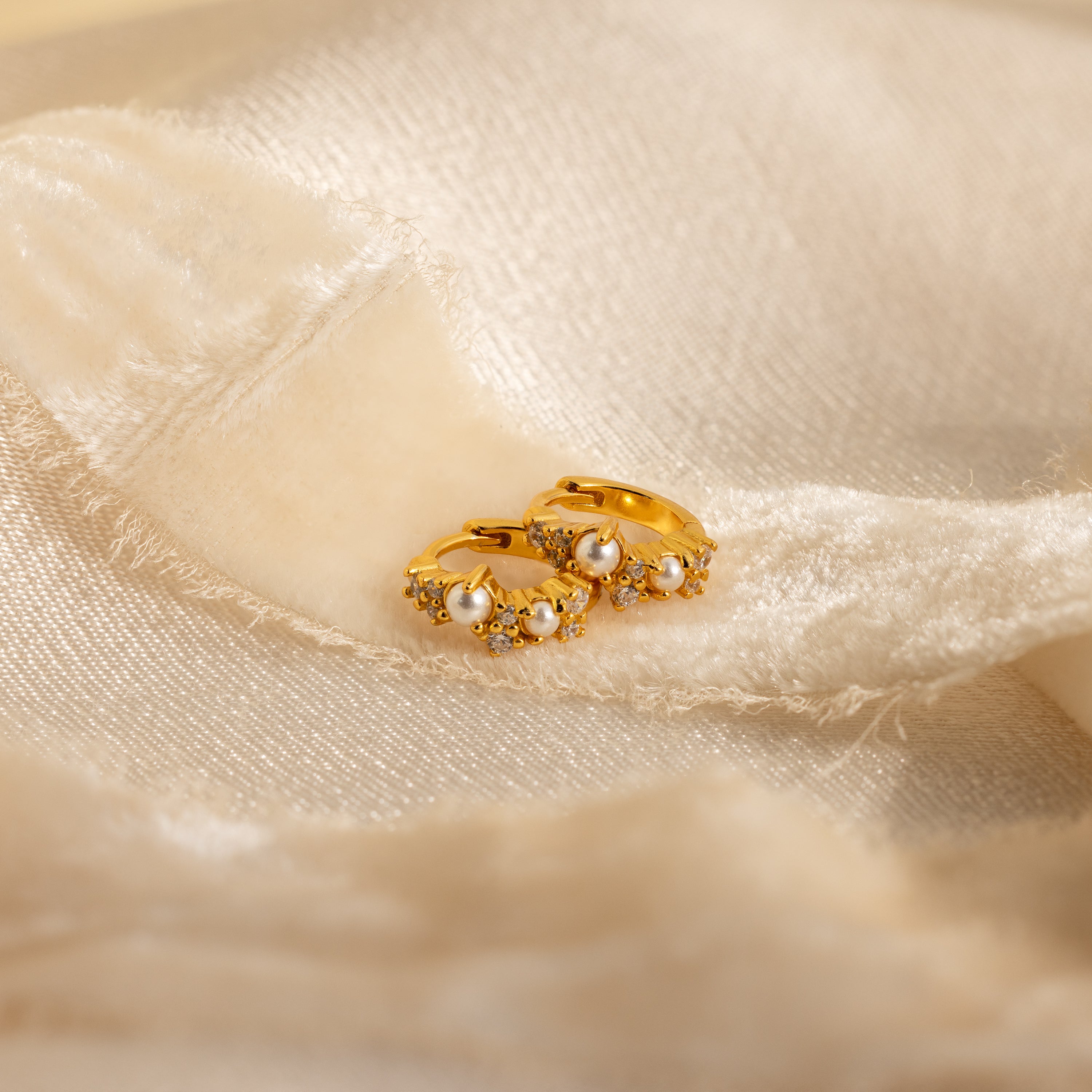 The Pearl Diamond Cluster Huggies in 18K Gold, featuring pearls and diamond accents, are displayed on soft, cream-colored fabric.