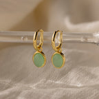 Jade Drop Hoops feature round jade gemstones hanging from huggie earrings, elegantly displayed on a clear rod with a soft beige fabric background.