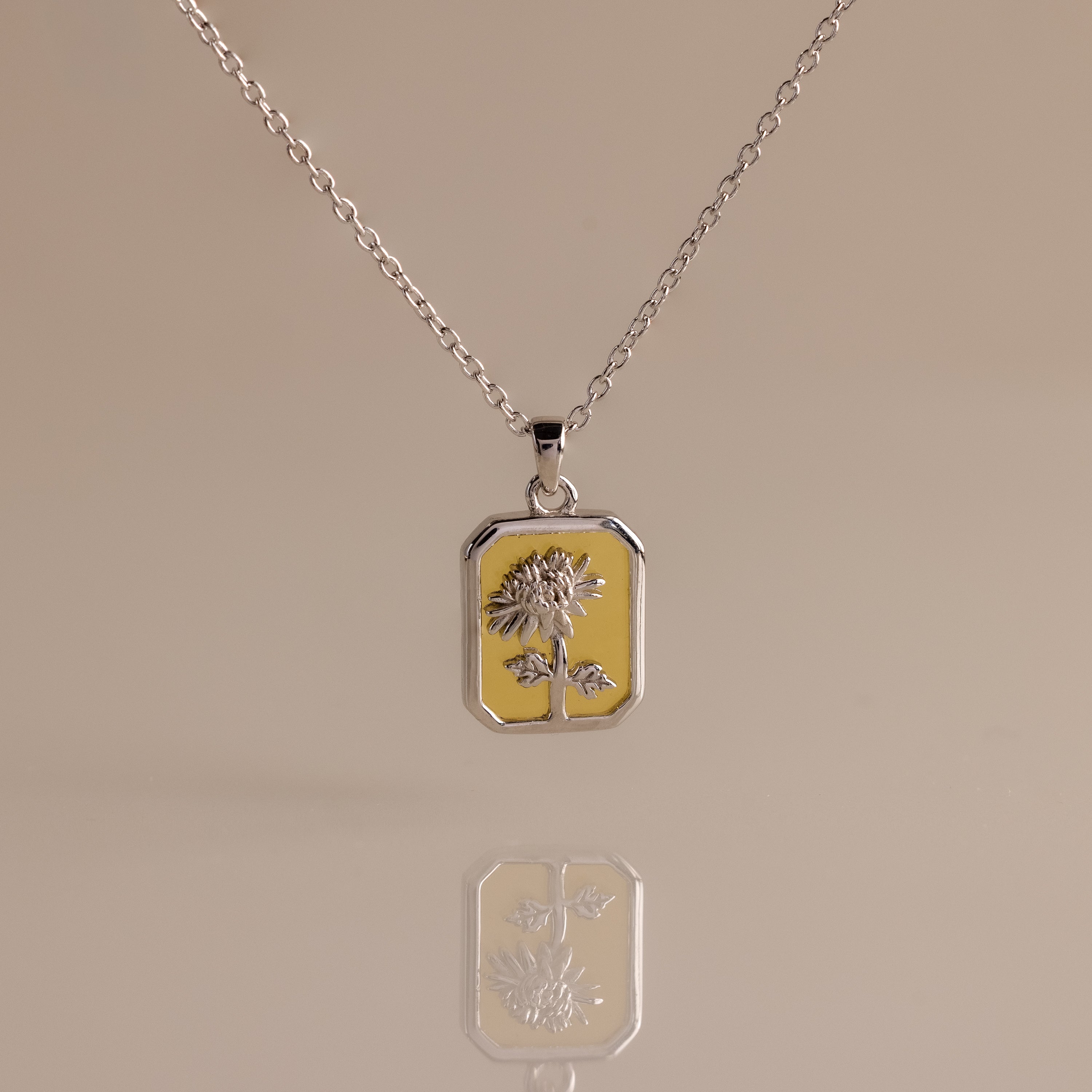 The Topaz Birth Flower Charm Necklace features a rectangular sunflower pendant, beautifully displayed on a beige background with an elegant reflection—ideal for those seeking a unique and delicate jewelry statement.