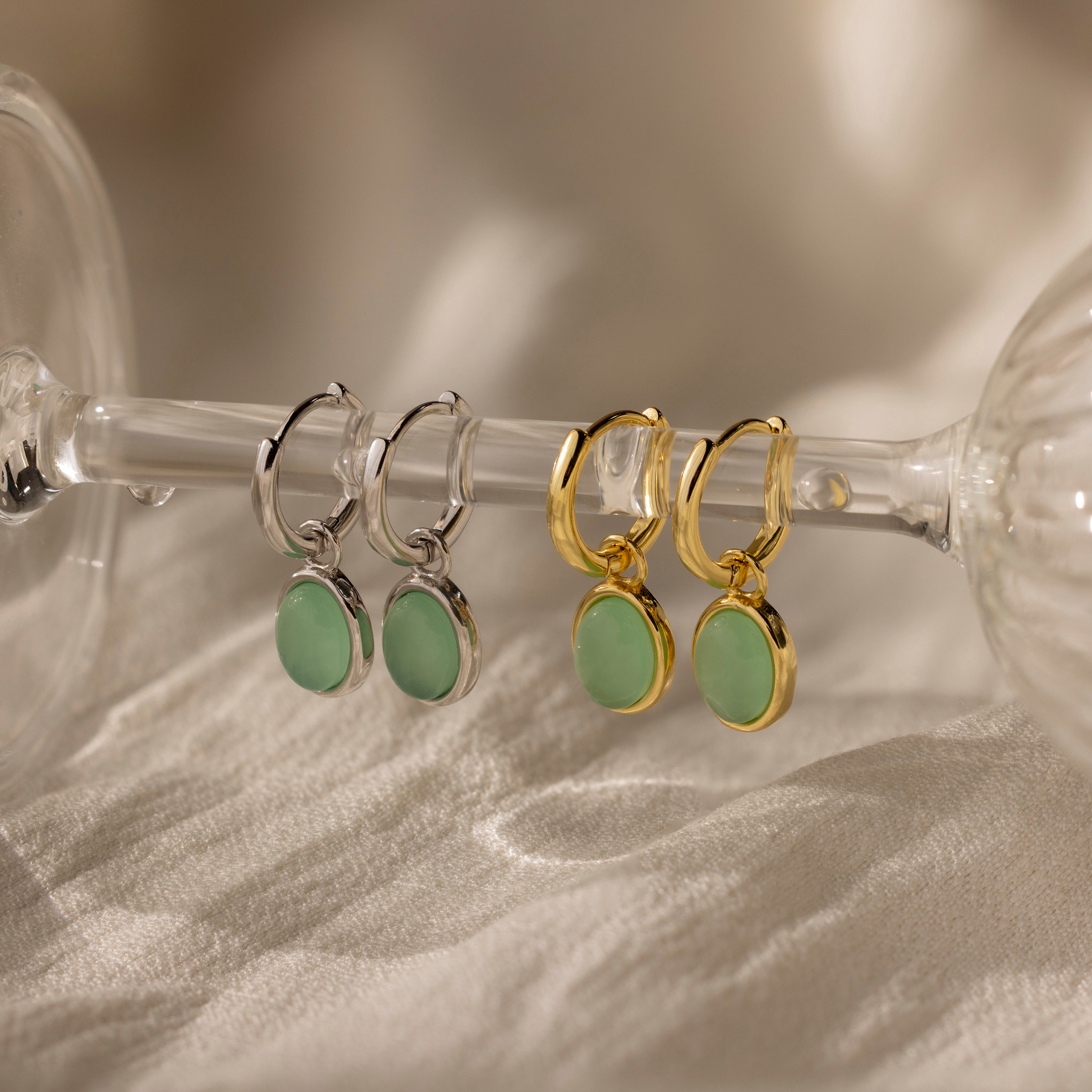 A pair of Jade Drop Hoops in Silver with green jade gemstone pendants is displayed on a glass stem.