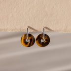 Tiger's Eye Duo Hoops in White Gold, featuring round brown tortoiseshell pendants inspired by golden-brown jewelry, displayed on a textured beige background.