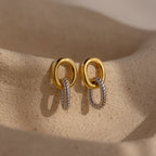 The Twisted Link Earrings in Mixed Metal rest on a textured beige surface with soft lighting, highlighting their sculptural artistry and modern elegance.