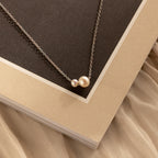 The Dainty Duo Pearl Necklace in Silver features two pearl pendants, shown on a beige and brown book—an elegant choice for weddings or a thoughtful gift for loved ones.