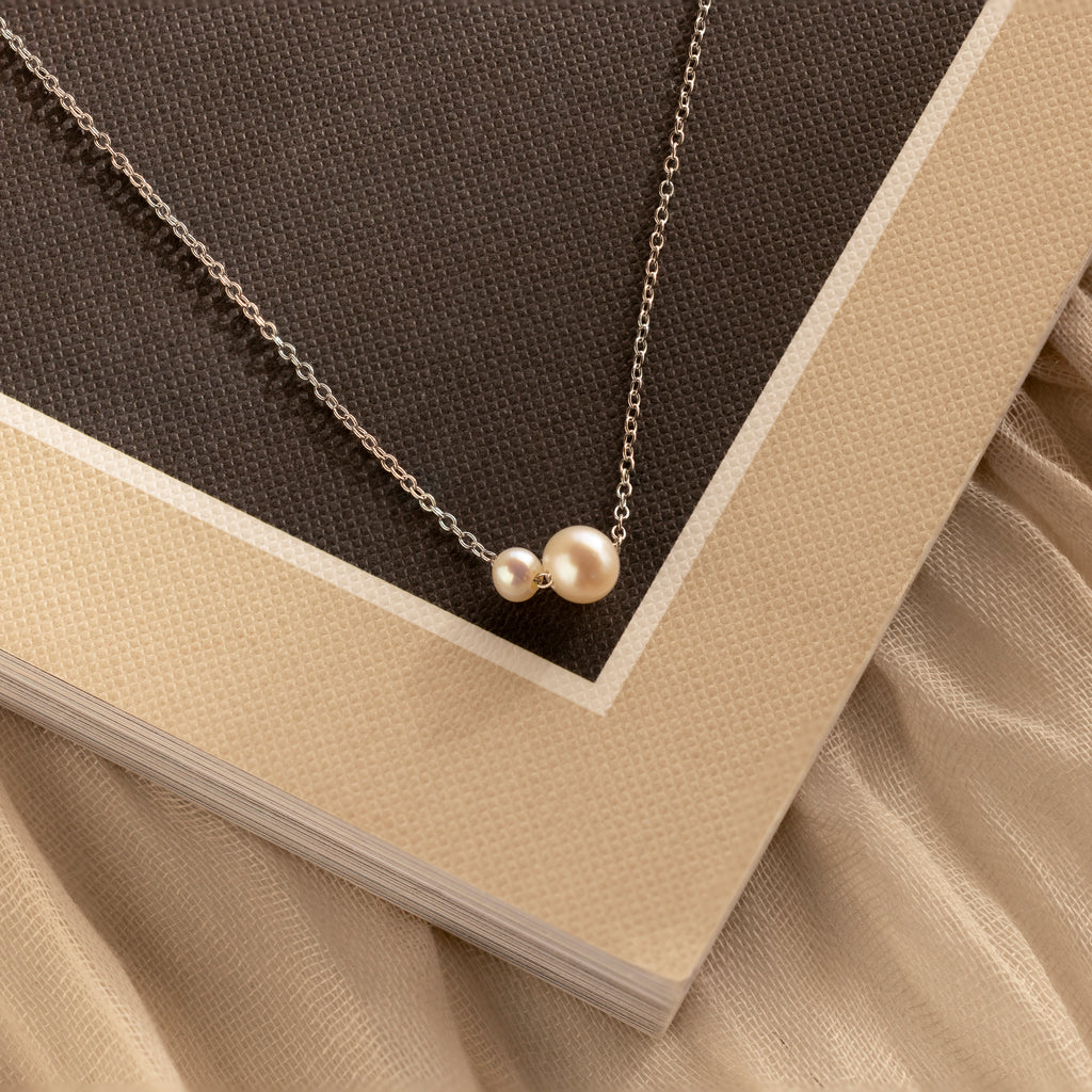 The Dainty Duo Pearl Necklace, featuring two pearls, rests on a textured brown and beige book—an elegant choice for wedding jewelry.