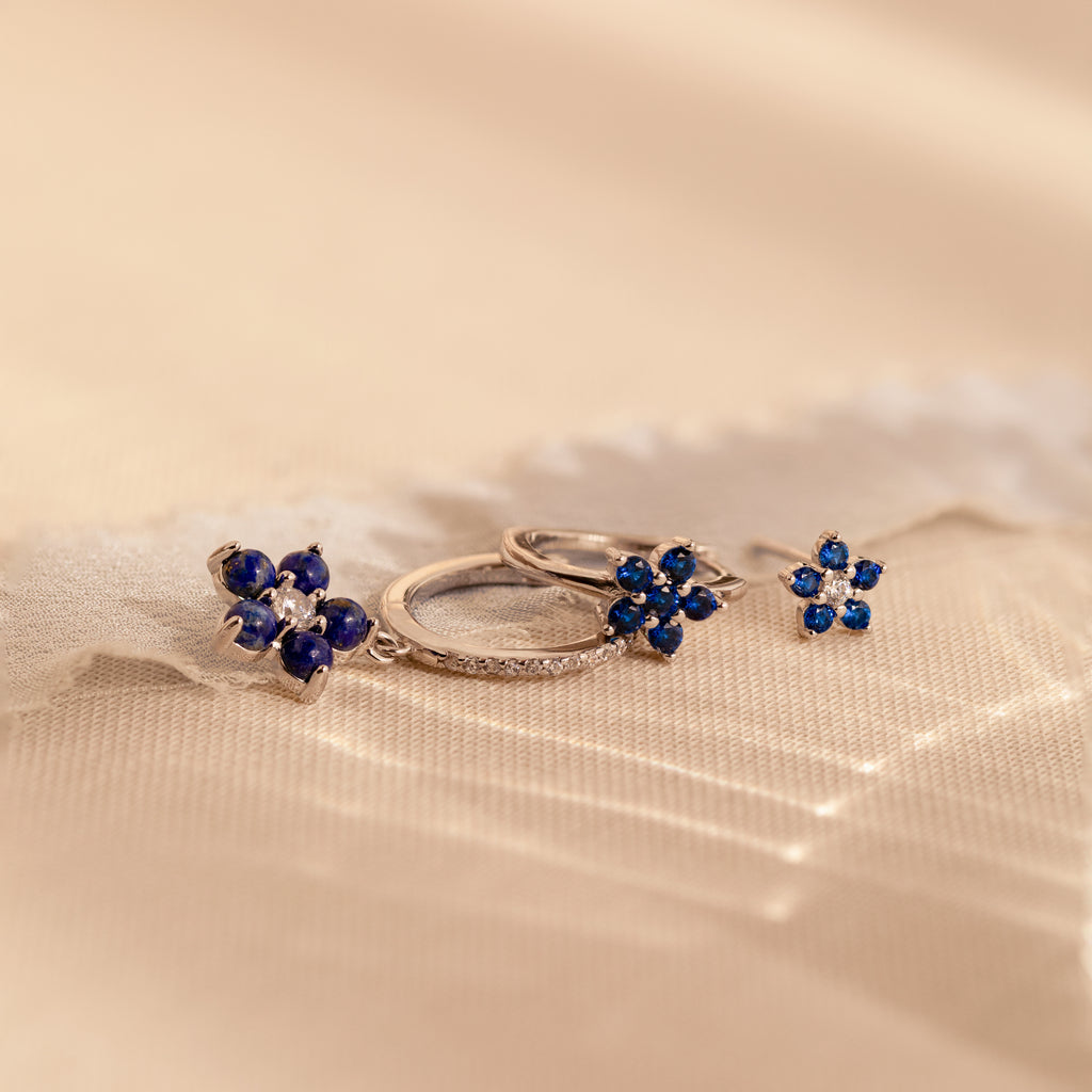 The Sapphire Flower Earrings Set - Silver features two pairs of silver earrings with blue gemstone flower designs on a soft, beige textured background—an elegant anniversary gift.