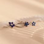 The Sapphire Flower Earrings Set - Silver features two pairs of silver earrings with blue gemstone flower designs on a soft, beige textured background—an elegant anniversary gift.