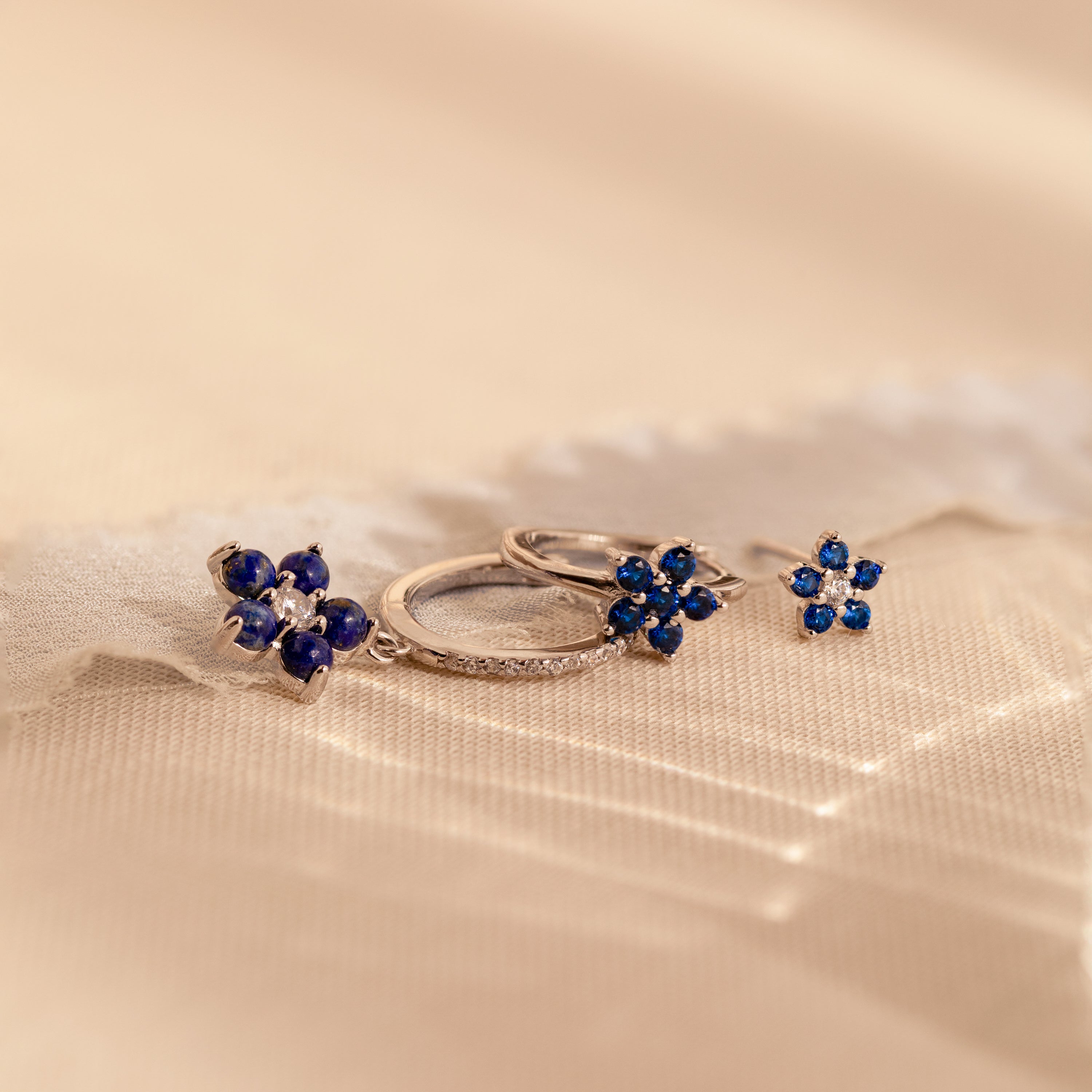 The Sapphire Flower Earrings Set - Silver features two pairs of silver earrings with blue gemstone flower designs on a soft, beige textured background—an elegant anniversary gift.