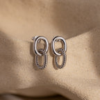 Twisted Link Earrings in White Gold, featuring smooth and textured chains, rest on a soft beige sandy background—modern earrings that embody timeless elegance.