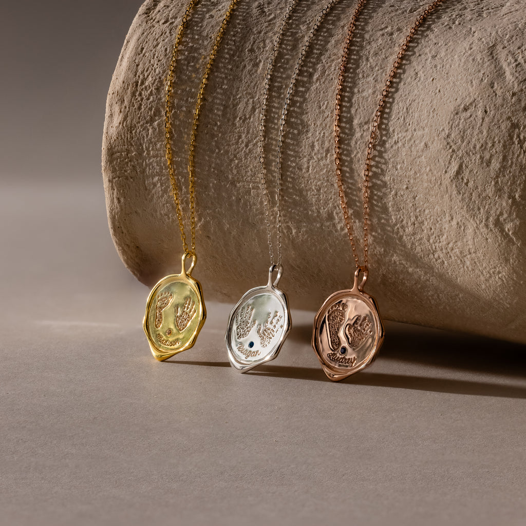 The Baby Hand & Footprint Necklace in Sterling Silver features an oval pendant on a textured chain, displayed on a stone surface—a timeless, personalized keepsake perfect for cherishing your baby's special moments.
