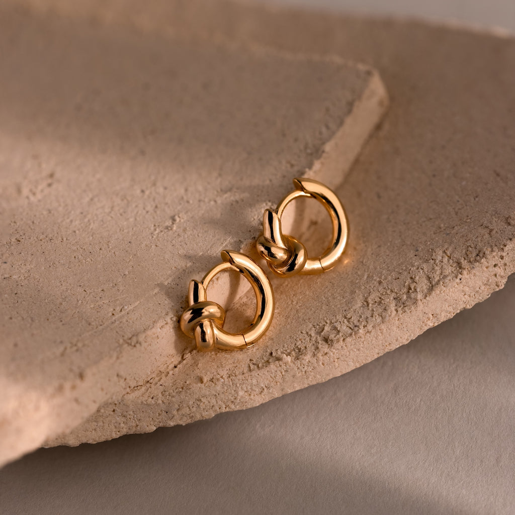 Love Knot Huggies in 18K Gold, featuring knot details that symbolize lasting bonds, elegantly displayed on a textured beige stone surface.