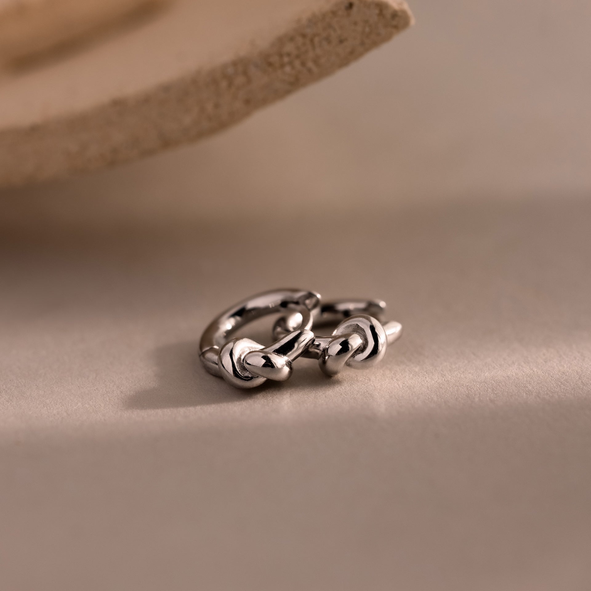 The Love Knot Huggies, a pair of silver hoop earrings, rest on a neutral surface with a textured accent above, symbolizing lasting bonds and making a meaningful gift.