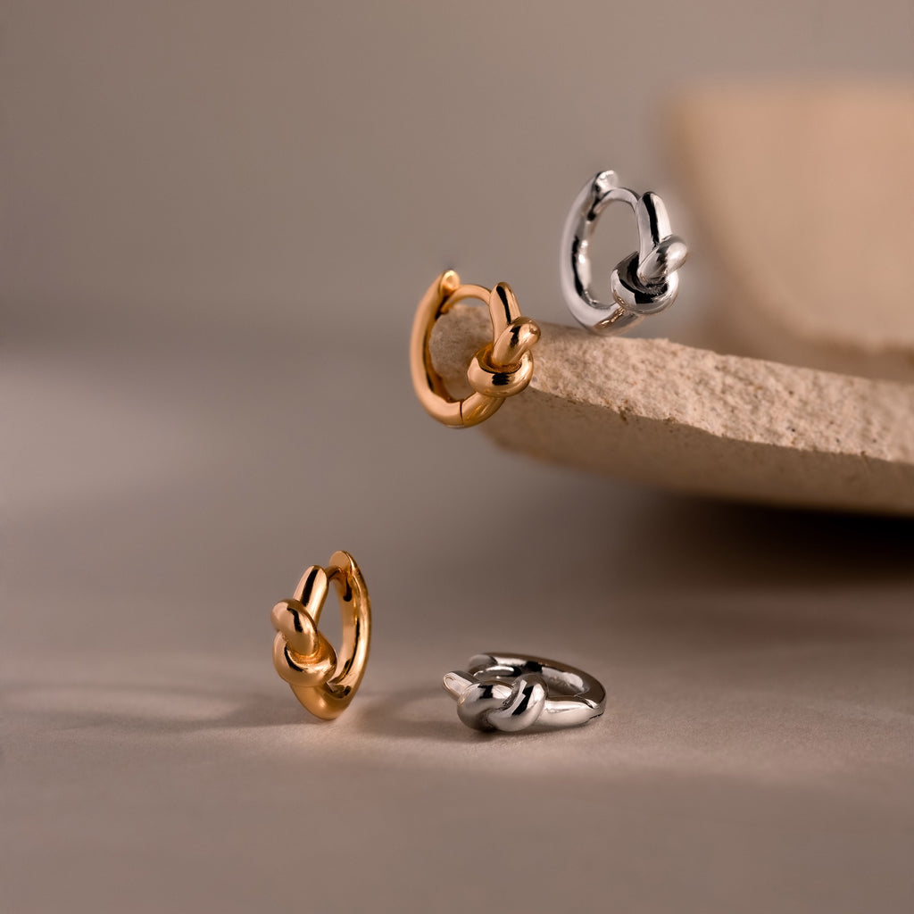 Two pairs of small hoop earrings, including the gold and silver Love Knot Huggies—ideal as a meaningful gift—displayed on a neutral textured surface.