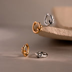 Two pairs of small hoop earrings, including the gold and silver Love Knot Huggies—ideal as a meaningful gift—displayed on a neutral textured surface.