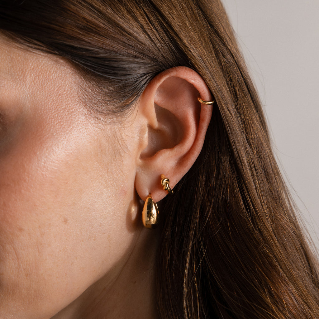 Close-up of a woman’s ear with three gold earrings, including Love Knot Huggies in 18K Gold—an elegant gift symbolizing lasting bonds, her brown hair tucked behind her ear.